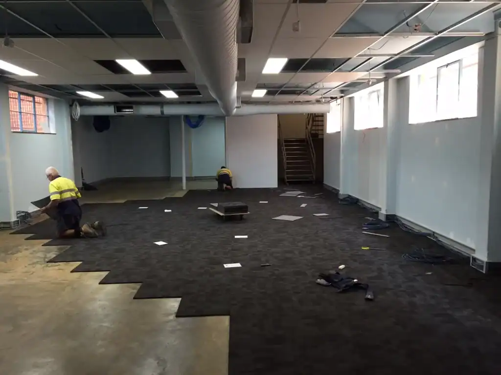 New office carpet installation site, workers laying down dark carpet tiles, construction in progress, bright window lighting, modern office renovation, 21st Century Business Equipment relevance.