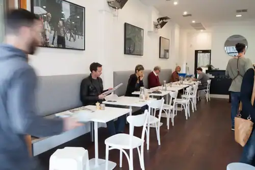 A bustling cafe interior with customers seated at tables and some walking, creating a lively atmosphere.