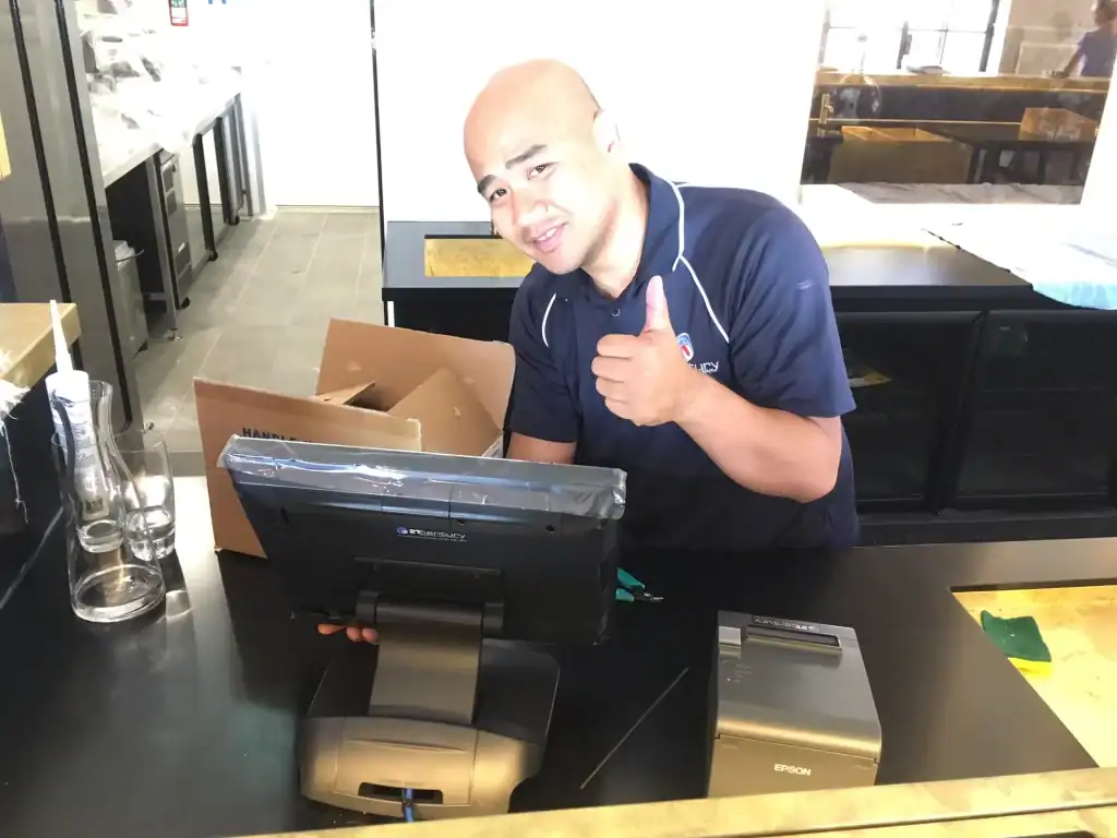 High-energy employee standing at checkout counter, giving thumbs-up and smiling with retail equipment.