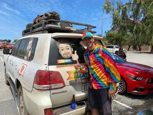A man standing next to a white station wagon with luggage on top, smiling and giving a thumbs-up.
