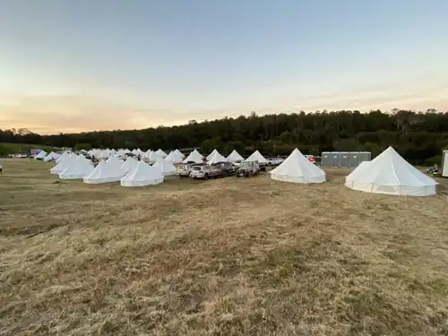 Temporary outdoor event tents and vehicles on a wide open field at sunset.