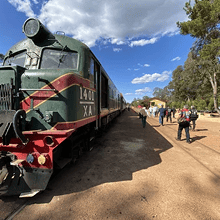 Steam locomotive train at a historical railway station, with people exploring and enjoying the scenic outdoor setting.
