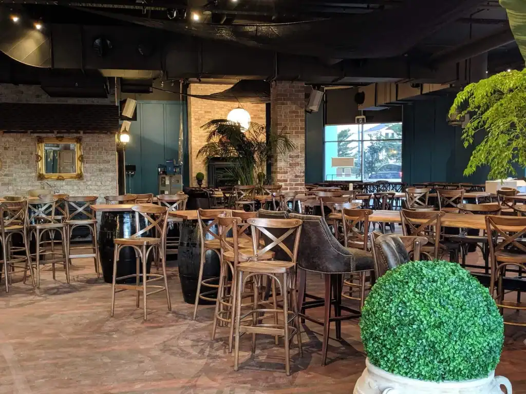 Modern restaurant interior with wooden chairs and potted plants, natural light through large windows, cozy atmosphere.
