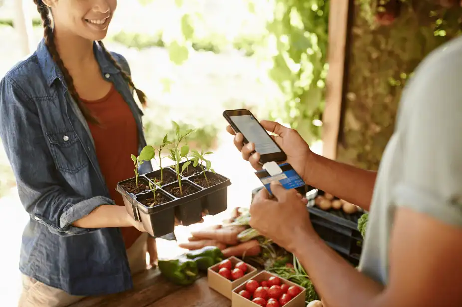 Greenhouse plant shop with people taking notes, holding plants and using smartphone for gardening assistance, outdoor farm market scene.