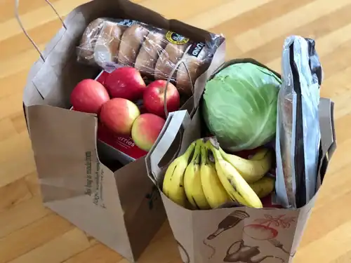Fresh produce grocery bags filled with apples, bananas, cabbage, bread, and packaged snacks on a hardwood floor.