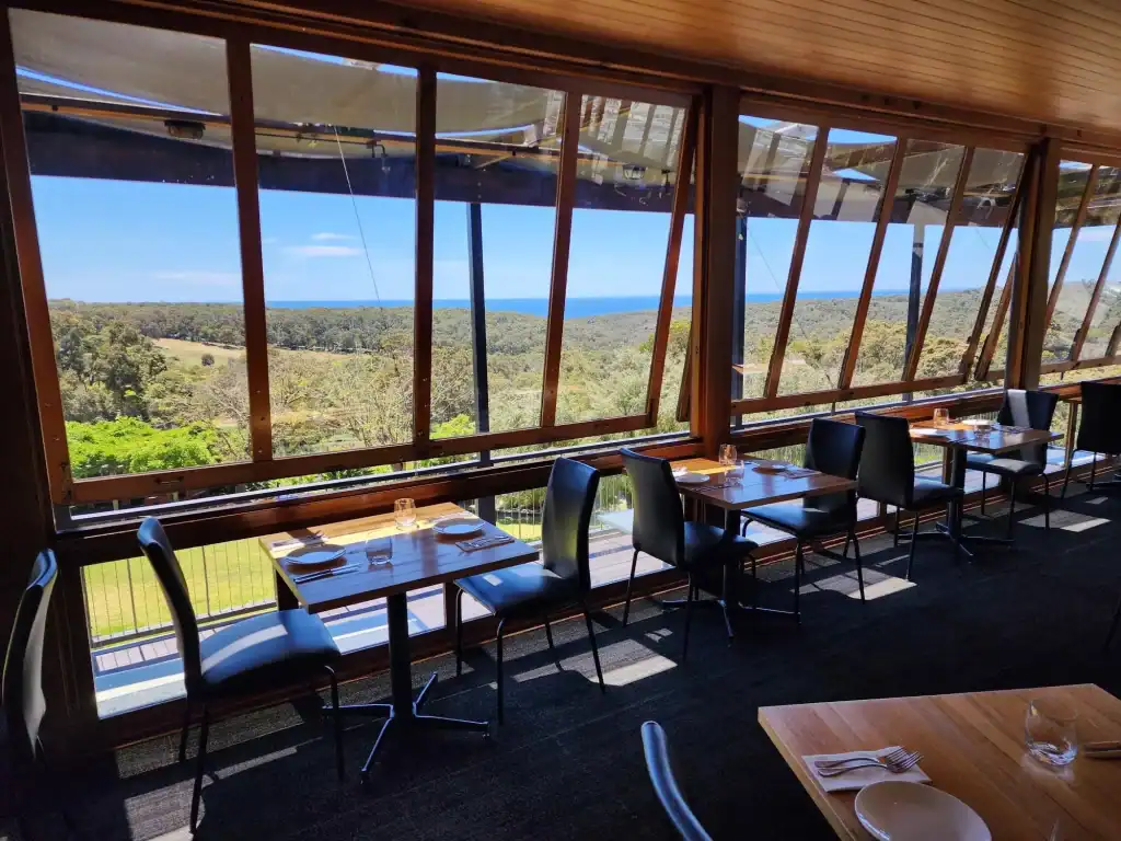 Scenic restaurant with large windows overlooking lush greenery and ocean view, elegant dining setup with black chairs and wooden tables.