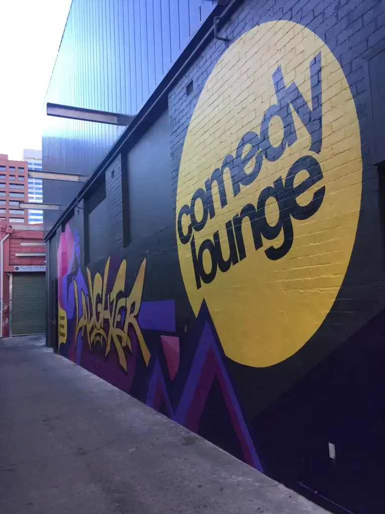 Colorful graffiti mural featuring the "Compong Lounge" logo on a brick wall, with buildings in the background.