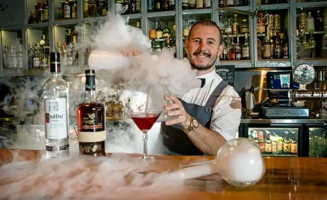 Barman creating a smoky cocktail at a bar with various spirits in the background.