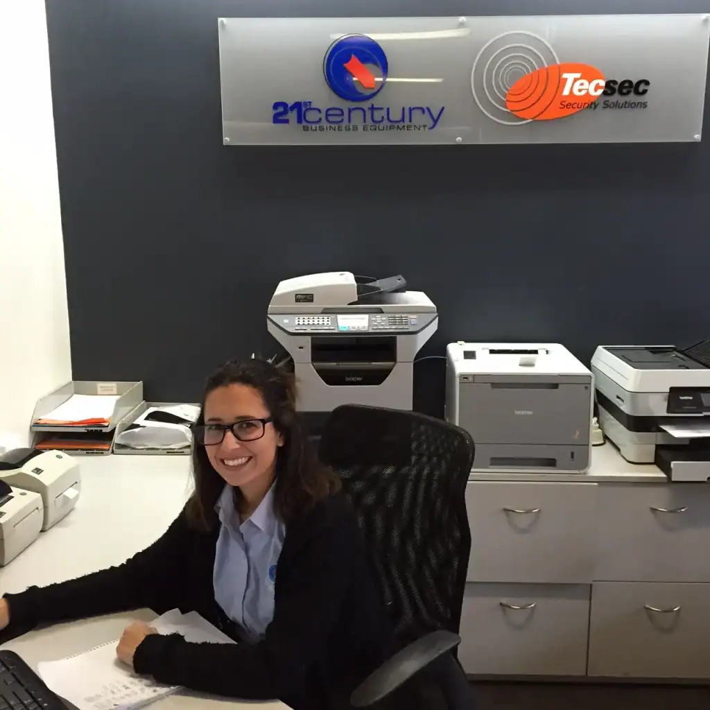 Professional woman at desk with office printers and photocopiers, representing 21st Century Business Equipment services.