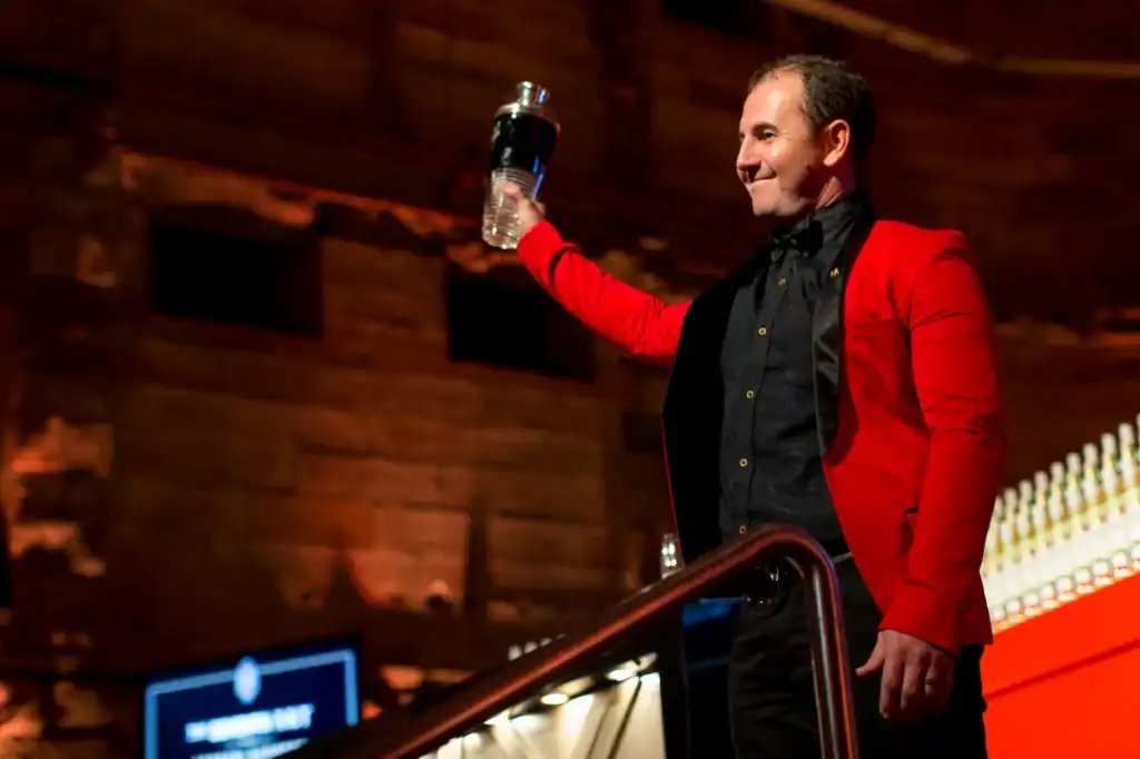 A man in a red blazer holding a bottle of water, smiling, at an indoor event with wooden walls and bar lights.