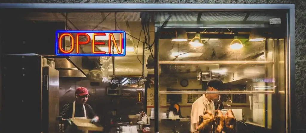 Neon "OPEN" sign in restaurant window, kitchen with chefs preparing food at night.