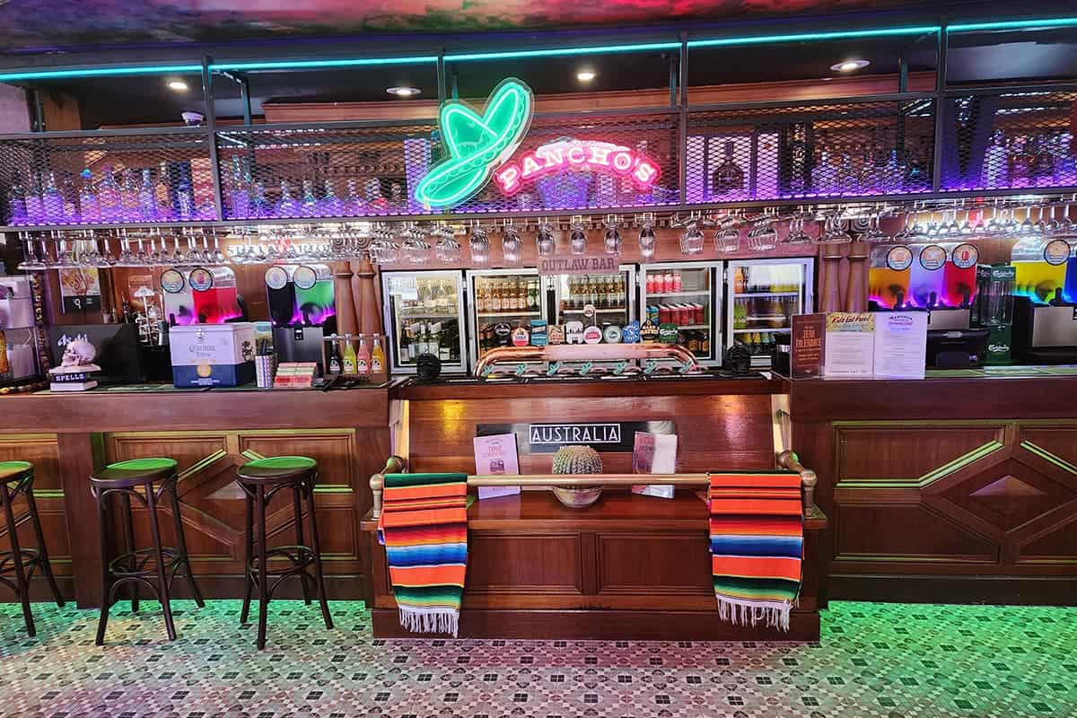 Neon sign at bar with colorful Mexican blankets and drinks in the background.