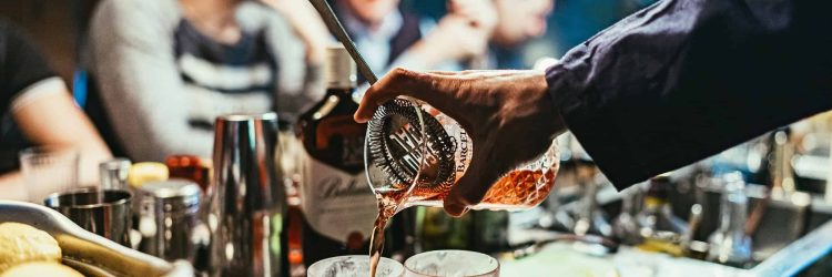 Cocktail being poured into glasses at bar counter in a lively social setting.