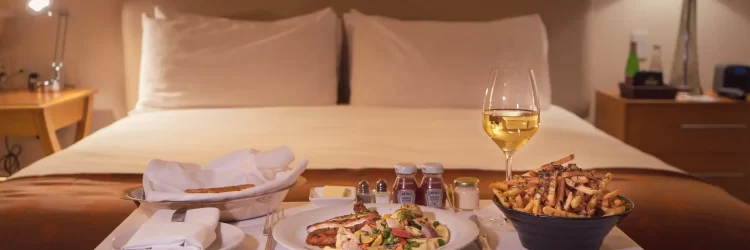Elegant hotel room with a bed and a table set for dining, including wine and food, in a cozy hospitality setting.